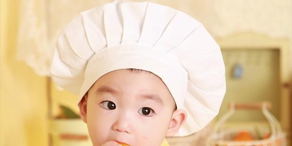 Un bambino con un cappello da chef mangia un'arancia, mostrando curiosità e allegria mentre esplora il mondo della cucina